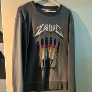 Grå tröja från Zadig & Voltaire - Säljer en grå tröja från Zadig & Voltaire i bra skick. Den har ett coolt tryck med texten 'ZADIG' och färgglada detaljer i rött, blått och gult. Perfekt för en avslappnad stil. Tröjan är långärmad och gjord i bomullsmaterial.