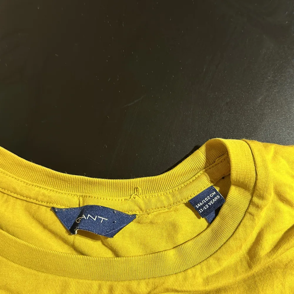 Säljer en snygg gul t-shirt från GANT med korta ärmar och ett coolt tryck på framsidan. Perfekt för sommardagar eller avslappnade tillfällen. T-shirten är i bra skick och gjord av mjukt bomullsmaterial.. T-paidat.