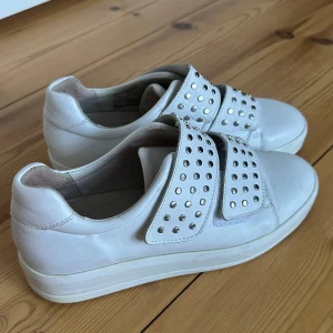 Vita sneakers med nitar - Säljer ett par vita sneakers i skinn med coola nitar på ovansidan. De har en stilren design utan snörning och är i bra skick. Perfekta för både vardag och fest! Storlek är inte angiven, men de ser ut att passa en normal passform. Storlek 39