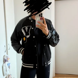 Svart bomberjacka med tryck från Bershka  - Säljer en svart bomberjacka med coola tryck och detaljer. Jackan har vita knappar och ett stort 'V' med en blomma på framsidan. Ärmarna har texten 'Authenticity' och är gjorda av ett skinnliknande material. Perfekt för höst och vår! Liten skada på axeln se bild 