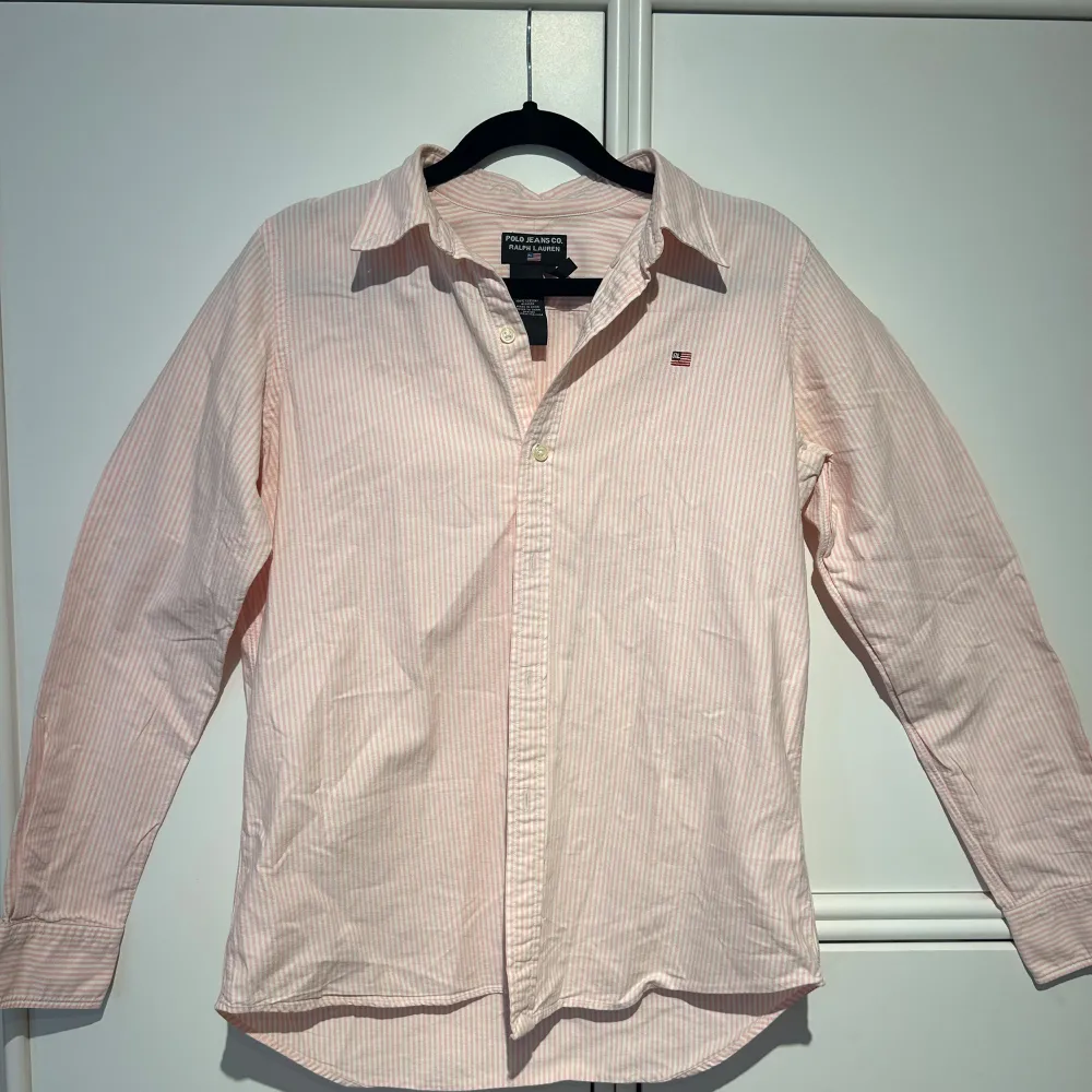 Säljer en snygg rosa och vit randig skjorta från Polo Jeans Co. av Ralph Lauren. Skjortan har långa ärmar och knappar framtill. Perfekt för både vardag och festliga tillfällen. Tror att det är herr men passar ju dam också därav unisex!. Skjortor.