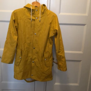 Gul regnjacka från 157 - Säljer en snygg gul regnjacka från 157 i storlek XS. Jackan har en praktisk huva och stängs med knappar. Den har två fickor framtill och är i mycket bra skick. 