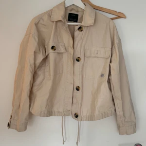 Beige jacka från Bershka - Säljer en snygg beige jacka från Bershka i storlek M. Jackan har långa ärmar och stora knappar framtill. Den är i bra skick och har en justerbar dragsko i nederkanten för en perfekt passform. Perfekt för vår och höst!