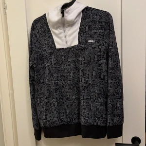 Svart och vit hoodie från BOSS - Säljer en snygg svart och vit hoodie från BOSS med ett coolt mönster av loggan över hela tröjan. Den har en dragkedja vid halsen och en stor vit huva. Perfekt för höst och vår när du vill vara både bekväm och stilren. Passar lika bra till vardags som till en avslappnad utekväll.