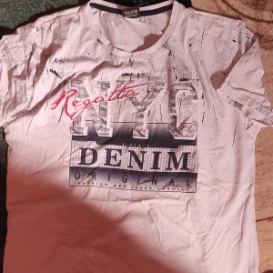 Vit t-shirt med tryck från Regatta - Säljer en vit t-shirt från Regatta med ett stort NYC Denim-tryck i grått och rött på framsidan. T-shirten är kortärmad och har en avslappnad stil, perfekt för vardagsbruk. Den är i bra skick och passar perfekt till jeans eller shorts. Den är köpt från Turkiet och är inte äkta