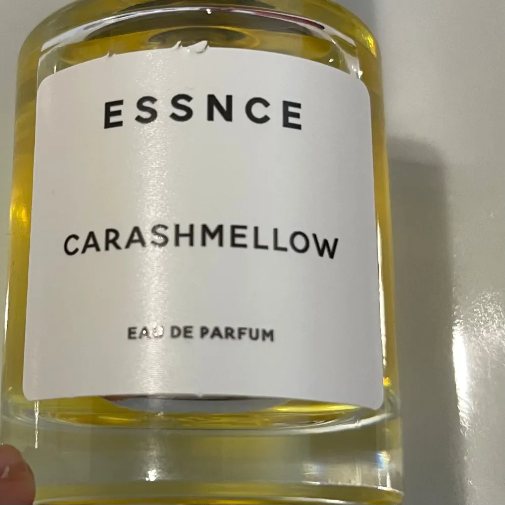 Säljer en flaska Essnce Carashmellow Eau de Parfum. Den har en stilren design med en genomskinlig glasflaska och svart kork. Parfymen är gyllengul och ger en lyxig känsla. Perfekt för dig som vill ha en unik doftupplevelse. Passar både till vardags och fest!. Parfym.