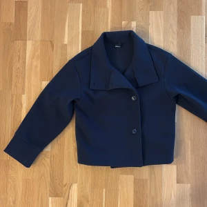 Mörkblå jacka från Gina Tricot - Säljer en stilren mörkblå jacka från Gina Tricot. Jackan har en klassisk krage och knäppning framtill. Perfekt för höst och vår, med en normal passform och långa ärmar. Den är i mycket bra skick och passar både till vardags och lite finare tillfällen.