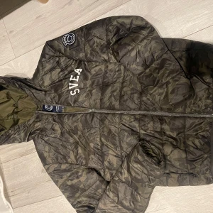 Camouflage pufferjacka från Svea - Säljer en snygg och varm camouflagefärgad pufferjacka från Svea. Jackan har en dragkedja framtill och en praktisk huva. Perfekt för höst och vinter! Den är i mycket bra skick och redo för nya äventyr.