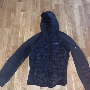 Svart pufferjacka från Peak Performance - Säljer en svart hybridjacka från Peak Performance i mycket bra skick. Jackan har en dragkedja framtill och en praktisk huva. Perfekt för kyliga dagar och passar både till vardags och utomhusaktiviteter. Storlek är inte angiven, men den ser ut att ha en normal passform.