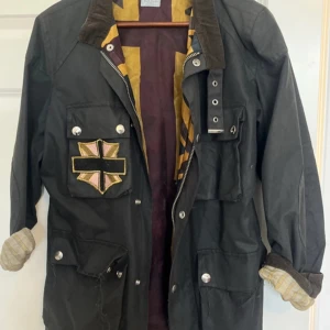 Svart jacka från Paul Smith - Säljer en cool svart jacka från Paul Smith, tillverkad i Italien. Den har långa ärmar och en unik broderad detalj på bröstet. Jackan har flera fickor med knappar och en dragkedja framtill. Perfekt för höst och vår!