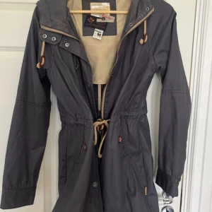 Grå parkajacka från Khujo - Säljer en snygg grå parkajacka från Khujo i storlek M. Jackan har långa ärmar och en praktisk huva med dragsko. Den har dragkedja och knappar framtill samt justerbar midja för perfekt passform. Perfekt för höst och vår!