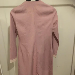 Rosa långärmad kappa med broderi - Säljer en söt rosa klänning med långa ärmar och vackert blombroderi på bröstet. Klänningen har en elegant design med en dekorativ knäppning framtill. Perfekt för vår och sommar!