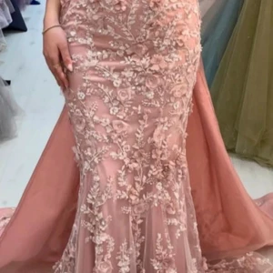 Rosa långklänning med spetsdetaljer - Fantastisk rosa långklänning med vackra spetsdetaljer och blommönster. Klänningen har en elegant slits och är perfekt för festliga tillfällen. Materialet är troligtvis satin vilket ger en lyxig känsla. Passformen är tight och framhäver figuren på ett smickrande sätt.