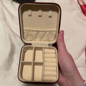 Röd smyckeskrin med dragkedja - Säljer ett snyggt och praktiskt rött smyckeskrin med dragkedja. Insidan är klädd i mjukt beige tyg och har flera fack för att organisera dina smycken. Perfekt för att hålla ordning på ringar, halsband och örhängen när du reser eller hemma.