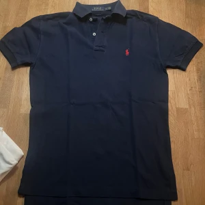 Mörkblå pikétröja från Ralph Lauren - Snygg mörkblå pikétröja från Ralph Lauren med klassisk krage och knappar. Den har en röd broderad logotyp på bröstet. Perfekt för en stilren och avslappnad look. Pikén är i bra skick och är i storlek XS