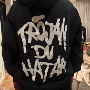 Norlie & kkv merch  - Säljer en svart hoodie med texten 'TRÖJAN DU HATAR' i vitt på ryggen. Den har en klassisk design med huva och en stor ficka framtill. Perfekt för en avslappnad stil. Köpt på konsert av Norlie & kkv! Större i storleken❤️