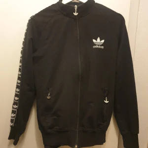 Svart tracksuit från Adidas - Snyggt svart sett från Adidas med dragkedja och ribbade muddar. Jackan har Adidas-logga på bröstet och längs ärmarna. Byxorna har elastisk midja med snörning och logga längs sidorna. Perfekt för en sportig look! 