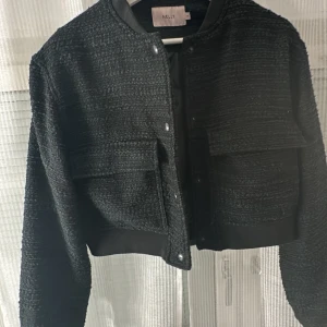 Svart croppad jacka från Nelly - Snygg svart croppad jacka från Nelly med knappar framtill. Jackan har en texturerad yta och två fickor på bröstet. Perfekt för en stilren look.