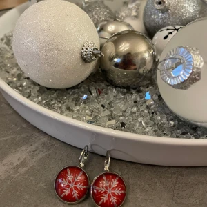 Röda örhängen med snöflingemotiv - Snygga röda örhängen med ett vitt snöflingemotiv, perfekta för vintersäsongen. De har en rund form och en glansig yta som ger en festlig känsla. Passar utmärkt till jul och nyårsfiranden.