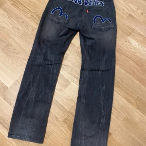 Svarta jeans från Evisu size 28~31 - Snygga svarta jeans från Evisu med unika blå broderade detaljer på bakfickorna och logotypen på baksidan. Jeansen har en klassisk passform med raka ben och normal midja. Perfekta för dig som vill ha en stilren men ändå iögonfallande look.