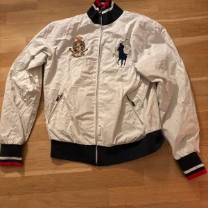 Vit jacka från Polo Ralph Lauren - Säljer en stilren vit jacka från Polo Ralph Lauren med broderad logga och emblem på bröstet. Jackan har dragkedja och ribbade muddar i svart, rött och vitt. Perfekt för en sportig look! HAR STORLEK L MEN KÄNNS SOM M