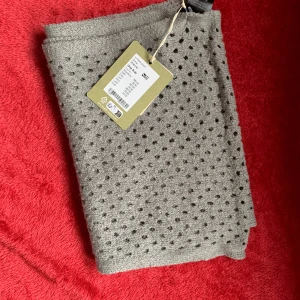Grå halsduk från Gudrun Sjödén  - Snygg beige halsduk från Gudrun Sjödén med ett diskret hålmönster. Perfekt för att hålla dig varm och stilren under kyliga dagar. Tillverkad med omtanke om miljön.