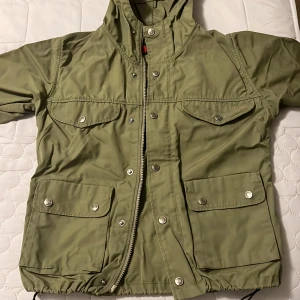 Grön jacka från Fjällräven - Säljer en grön jacka från Fjällräven i storlek XXS. Jackan har en praktisk huva och flera fickor med knappar. Perfekt för utomhusaktiviteter med sin robusta design och dragkedja framtill.