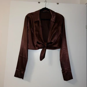 Brun satinblus från NLY One - Snygg brun satinblus från NLY One med knyt framtill. Blusen har långa ärmar och en klassisk krage, perfekt för en chic look.
