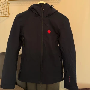 Skidjacka från J.Lindeberg - Snygg skidjacka från J.Lindeberg med huva och dragkedja. Den har en röd logotypdetalj på bröstet och märkesnamnet på huvan. Perfekt för kyligare dagar. Säljer matchande skidbyxor