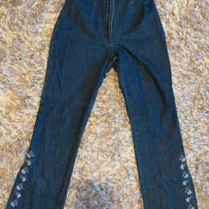 Blå jeans med broderade blommor från Brooker - Snygga blå jeans från Brooker med broderade blommor längs benen. Har split i benen och är korta för mig som är 168cm. Använd få gånger!