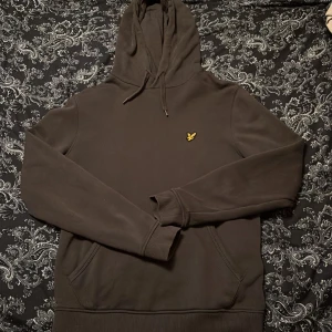 Hoodie från Lyle & Scott - Säljer en stilren grå hoodie från Lyle & Scott med deras ikoniska logga på bröstet. Tröjan har en praktisk känguruficka och justerbar huva med dragsko. Perfekt för en avslappnad look. Fint skick och den säljs pågrund av den blivit för liten. Hör av er vi frågor🤩