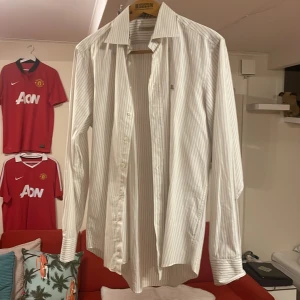 Vit randig skjorta från Burberry  - Snygg vit skjorta från Burberry Brit med tunna grå ränder och broderad logga på bröstet. Skjortan har en klassisk krage och knappar framtill. Perfekt för en stilren look. Den är ett par år gammal men har knappt används så väldigt bra skick!