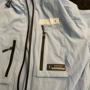 Ljusblå vindjacka från Hoodrich - Snygg ljusblå vindjacka från Hoodrich med dragkedja och flera praktiska fickor. Jackan har en justerbar huva och stilrena svarta detaljer. Perfekt för blåsiga dagar.