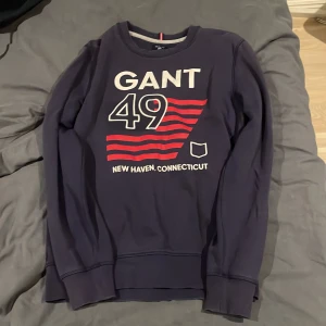 Mörkblå sweatshirt från GANT - Säljer en mörkblå sweatshirt från GANT med tryck i rött och vitt. Tröjan har rund hals och långa ärmar. Perfekt för en avslappnad stil. 7/10 i skick då den är använd rätt mycket men har inga hål eller liknande skavanker.