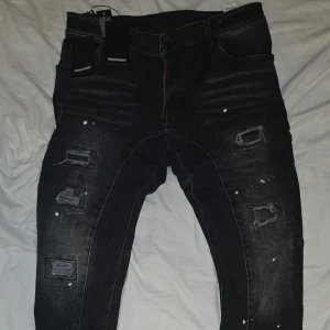 Svarta jeans med slitningar från Dsquared2 - Säljer helt nya snygga svarta jeans från Dsquared2 med coola slitningar och färgstänk. De har en modern design med knappar och dragkedja. Perfekta för en trendig look.