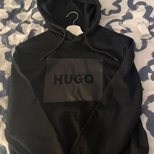 Svart hoodie Hugo boss - Säljer en stilren svart hoodie från Hugo med en stor logga på framsidan. Tröjan har en klassisk design med huva och dragsnören, perfekt för en avslappnad look. Perfekt skick, inga hål eller färgskador.