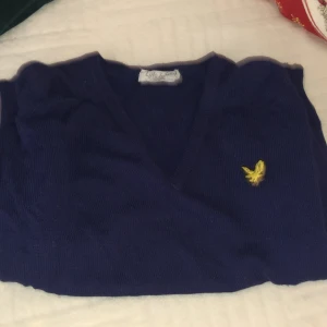 Mörkblå tröja från Lyle & Scott - Säljer en mörkblå tröja från Lyle & Scott med en gul logotyp på bröstet. Tröjan har en v-ringad design och är tillverkad i 100% akryl. Perfekt för en stilren och klassisk look.