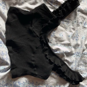 Svart ribbad topp från SHEIN - Snygg svart ribbad topp från SHEIN med volangdetaljer på axelbanden. Toppen har en oneshoulder-design 