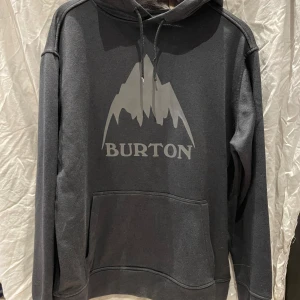 Burton Hoodie - Säljer en snygg grå hoodie från Burton med deras logga  tryckt på framsidan. Hoodien har en klassisk känguruficka och justerbar dragsko i huvan. Perfekt för kyliga dagar eller att ha under skidjackan. Storlek S men sitter normalt på mig som bär M - L
