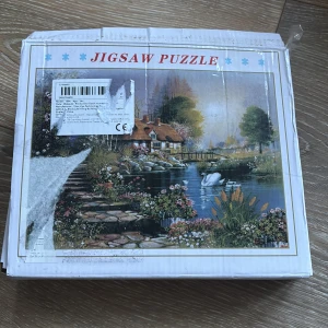 Pussel med landskapsmotiv - Ett vackert pussel med ett idylliskt landskapsmotiv som visar en stuga vid en sjö med svanar och en stenlagd stig. Perfekt för pusselentusiaster som gillar natur och avkopplande motiv. Pusslet är använt endast en gång och är helt komplet, ingen bit saknas. Förpackningen är trasig men pusselbitarna är hela. 