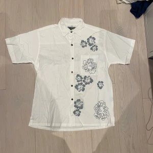 Flysurf skjorta blommor - Snygg vit skjorta från Flysurf med korta ärmar och ett stilrent blommönster i blått. Skjortan har bruna trä-knappar framtill och en krage. Skulle behöva en strykning annats helt felfri. Blommigt tryck både fram och bak. Riktigt somrig skjorta