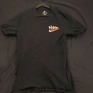 Svart t-shirt från Nike - Snygg svart t-shirt från Nike med ett coolt tryck i rött och vitt på både fram- och baksidan. Perfekt för en avslappnad stil. Tillverkad i mjukt material för extra komfort.