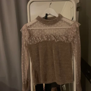 Beige tröja med prickig mesh och volangdetaljer - Säljer en beige tröja med långärmad design och prickig mesh upptill. Tröjan har volangdetaljer vid axlarna och en hög krage. Perfekt för en stilren och unik look.