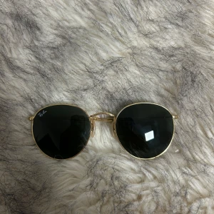 Runda solglasögon från Ray-Ban - Snygga runda solglasögon från Ray-Ban med guldfärgad metallram och mörka linseer. Använts i några månader. De har justerbara nässadlar 