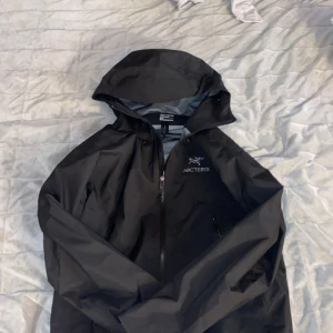 Svart vindjacka från Arc’teryx - Säljer en stilren svart vindjacka från Arcteryx. Jackan har en praktisk huva och dragkedja framtill. Perfekt för utomhusaktiviteter och skyddar mot vind och regn. Med dubbla inne fickor och en bra luva emot regn