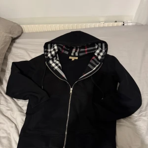 Svart Burberry zip - Snygg svart hoodie från Burberry med dragkedja och klassiskt rutigt foder i huvan. Perfekt för en stilren och bekväm look. Storlek M men hade passat bättre med någon med storlek S då den är kort i modell! 