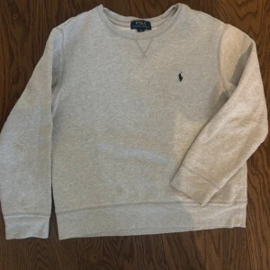 Grå sweatshirt från Polo Ralph Lauren - Säljer en klassisk grå sweatshirt från Polo Ralph Lauren med rund hals och det ikoniska logotypbroderiet på bröstet. Perfekt för en avslappnad stil. Storlek 152 barn 10/12