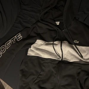 Svart och grå hoodie från Lacoste - Säljer en snygg svart och grå hoodie från Lacoste med dragkedja och huva. Den har en broderad krokodil på bröstet och texten 'LACOSTE' på ärmen. Perfekt för en avslappnad stil. Nypris 1700 och är använd ett fåtal gånger.