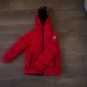 Röd dunjacka från Moncler - Snygg röd dunjacka från Moncler med huva och dragkedja. Jackan har ett broderat Moncler-märke på ärmen och praktiska fickor med dragkedjor. Perfekt för kyliga dagar.