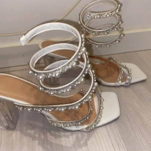 Vita sandaler med pärldetaljer - Eleganta vita sandaler med transparent klack och dekorativa pärlor. Sandalerna har en fyrkantig tå och flera remmar prydda med pärlor och glittrande stenar, vilket ger en lyxig känsla.